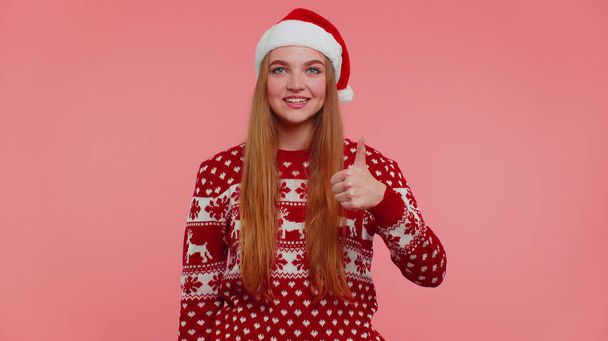 Chica divertida usa suéter rojo de Año Nuevo levanta pulgares hacia arriba está de acuerdo con algo bueno, como, fondo rosa - Foto, Imagen