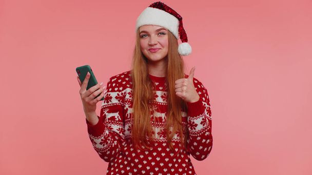 Woman in Christmas sweater looking smartphone display sincerely rejoicing win success luck, thumb up - Фото, зображення
