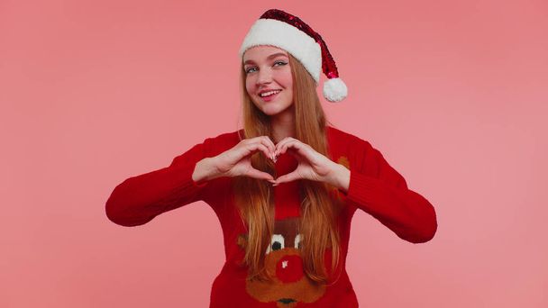 Smiling girl in Christmas sweater makes heart gesture demonstrates love sign expresses good feelings - 写真・画像
