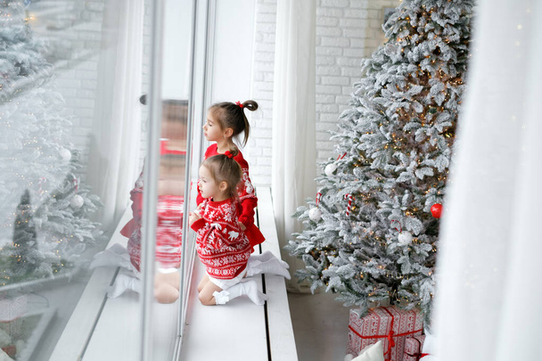 Los niños observan la nieve que cae fuera de la ventana, sentados en el alféizar de la ventana junto a un árbol de Navidad artificial decorado. Esperando milagros navideños. Hermanas en jerséis rojos de Navidad. - Foto, Imagen