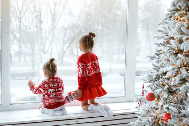 Две маленькие счастливые девочки смотрят первый снег через окно, сидя на подоконнике. Дети в рождественских красных свитерах рядом с украшенной елкой ждут канун Рождества. - Фото, изображение