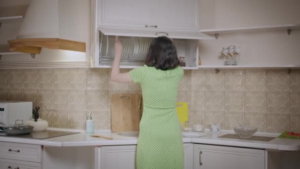 Menina morena em um vestido verde coloca as coisas em ordem no armário da cozinha, organiza os pratos - Filmagem, Vídeo