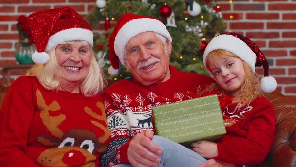 Senior-Großeltern mit Enkelin in Nikolausmütze feiern zu Hause am Weihnachtsbaum - Foto, Bild