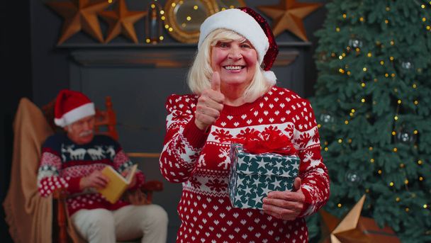Positive senior grandmother in Christmas sweater smiling friendly at camera and raises thumbs up - 写真・画像