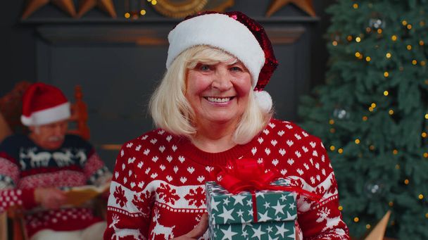 Idős nagymama ünnepi pulóverben, karácsonyi ajándékdobozt mutat be, mosolyog, kamerába néz - Fotó, kép