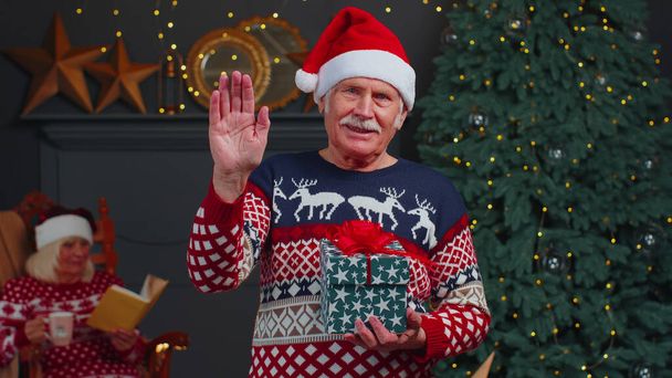 Ανώτερος παππούς σε χριστουγεννιάτικο πουλόβερ χαμογελώντας φιλικό στην κάμερα και χαιρετώντας τα χέρια gesturing γεια - Φωτογραφία, εικόνα