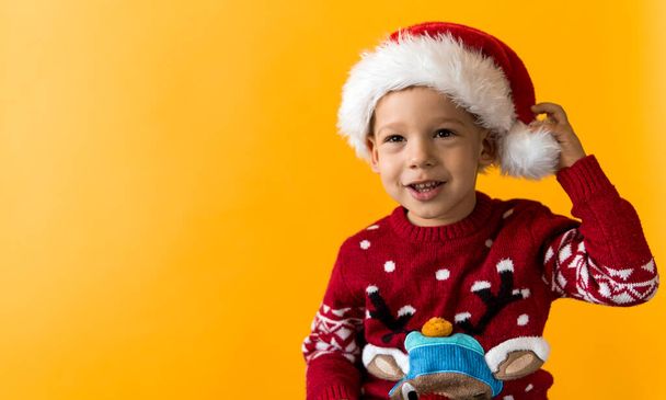 Portrait of happy smiling positive joyful positive preschool little boy in red warm santa hat showing thumb up on orange, yellow background. Winter, holiday, celebration, Christmas, New Year copy - Φωτογραφία, εικόνα