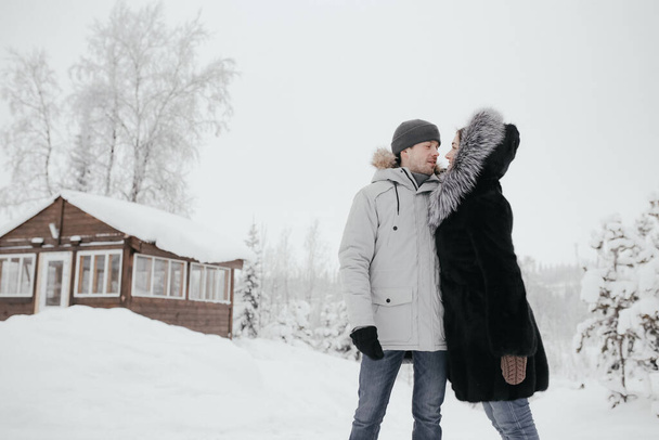 愛の美しいカップル、笑顔とお互いを見て、背景に小さな木造の家と雪 - 写真・画像