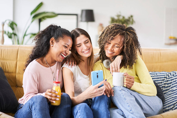 Grupo interracial de tres hermosas jóvenes amigas en casa riendo usando un teléfono inteligente para redes sociales - Foto, imagen