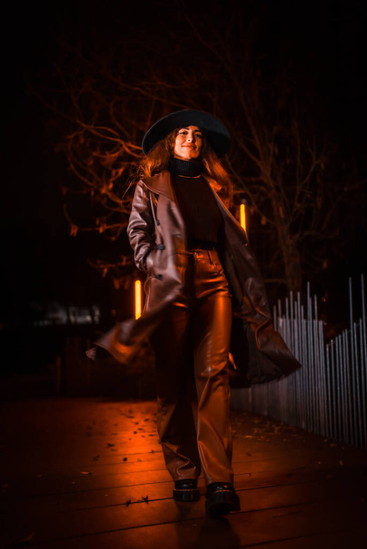 Girl in a hat in a portrait at night in a raincoat on a parcel. Caucasian brunette model looking at camera - Fotografie, Obrázek
