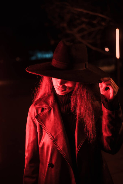 Girl in a hat in a portrait at night in a raincoat on a parcel. Illuminated with dim red light from the side and an out-of-focus tree - Photo, image