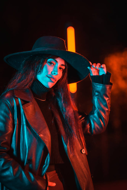 Caucasian brunette girl with hat at night in a park illuminated with blue light. Winter fashion and lifestyle at night, vertical photo - Fotografie, Obrázek