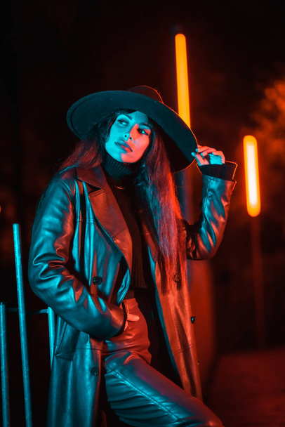 Caucasian brunette girl with hat at night in a park illuminated with blue light. Winter fashion and lifestyle at night, vertical photo - Fotó, kép