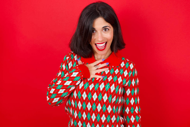 brunette caucasian girl wearing knitted sweater christmas over red background smiles toothily cannot believe eyes expresses good emotions and surprisement - Photo, Image