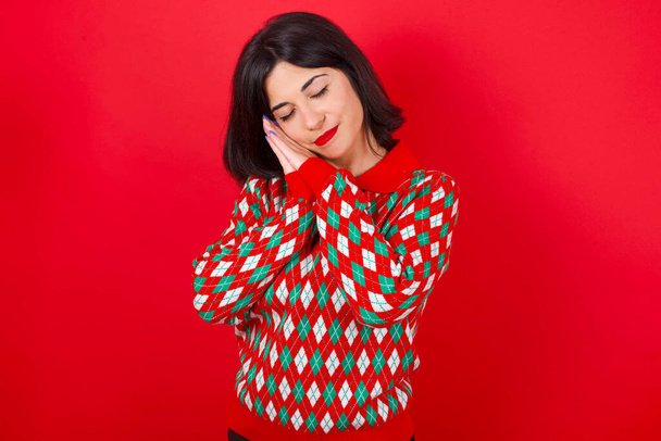 morena chica caucásica usando suéter de punto navidad sobre fondo rojo se apoya en palmas prensadas cierra los ojos y tiene agradables sueños sonrisa sobre algo - Foto, imagen
