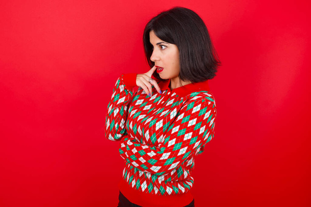 Astonished brunette caucasian girl wearing knitted sweater christmas over red background looks aside surprisingly with opened mouth. - Фото, изображение