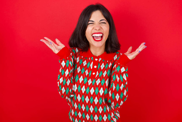 Pazzo indignato bruna caucasica ragazza indossa maglia maglione Natale su sfondo rosso urla forte e gesti urla rabbia furiosamente. Concetto di emozioni umane negative sentimenti - Foto, immagini