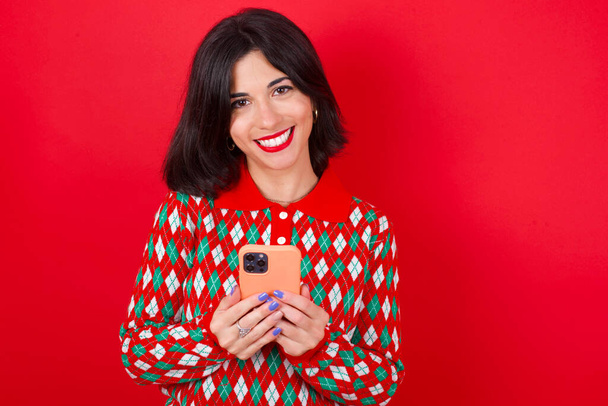 ブルネットの白人の女の子は赤い背景の上にニットセータークリスマスを身に着けていますモックアップコピースペース。携帯電話を使ってSMSメッセージを入力する - 写真・画像