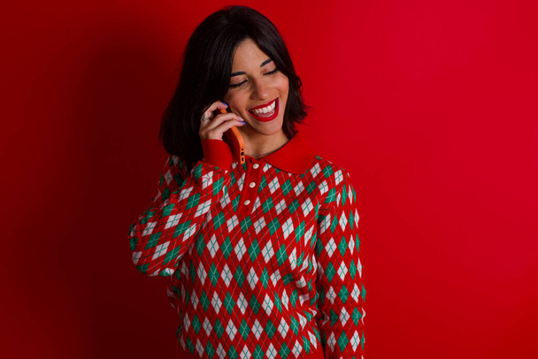 Agradable mirada morena feliz chica caucásica con suéter de punto navidad sobre fondo rojo tiene buena conversación telefónica y mira a un lado, tiene buen humor y sonríe positivamente mientras habla a través del teléfono celular - Foto, imagen