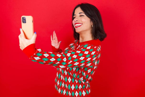 Brünettes kaukasisches Mädchen im Strickpullover vor rotem Hintergrund hält modernes Handy in der Hand und macht Videoanrufwellen in Hallo-Geste. Menschen modernes Technologiekonzept - Foto, Bild