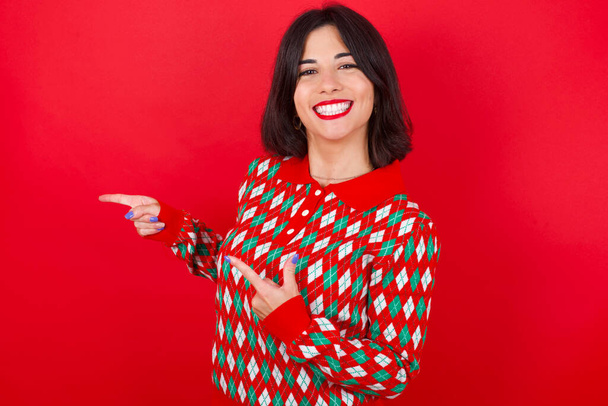 Optimistic brunette caucasian girl wearing knitted sweater christmas over red background points with both hands and  looking at empty space. - Fotografie, Obrázek