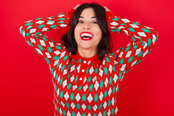 Veselé nadšené brunetky běloška dívka na sobě pletený svetr Vánoce nad červeným pozadím reaguje rostoucí ruce nad hlavou po obdržení skvělé zprávy. - Fotografie, Obrázek