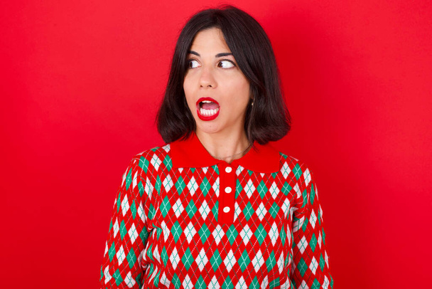 Sokkolt barna kaukázusi lány visel kötött pulóver karácsony felett piros háttér nézd üres tér nyitott szájjal sikoltozik: Ó, Istenem! Ezt nem hiszem el.. - Fotó, kép