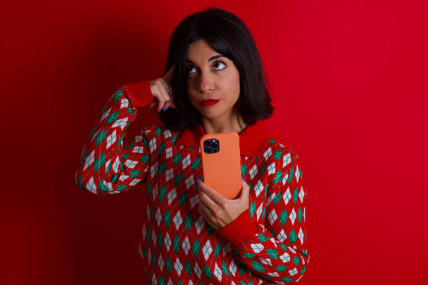 morena chica caucásica usando jersey de punto navidad sobre fondo rojo sosteniendo gadget mientras que sobresale lengua - Foto, Imagen