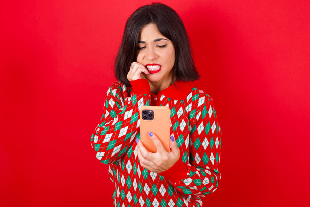 Porträt eines ziemlich verängstigten brünetten kaukasischen Mädchens mit Strickpullover Weihnachten auf rotem Hintergrund plaudert beißende Nägel, nachdem sie ein paar beängstigende Nachrichten auf ihrem Smartphone gelesen. - Foto, Bild
