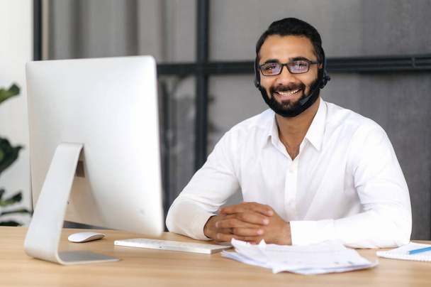 Porträt eines gutaussehenden positiven intelligenten indischen Mannes mit Brille und Headset, Experte, Berater oder Mentor, der freundlich lächelnd direkt in die Kamera blickt. Servicekonzept, Hotline - Foto, Bild