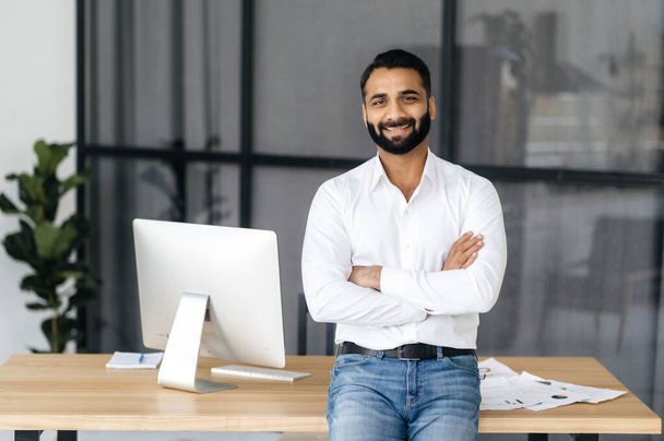Retrato de um homem indiano confiante de sucesso, mentor, gerente ou especialista em TI, vestindo uma camisa branca, de pé perto da mesa no escritório com os braços cruzados, olhando para a câmera e sorrindo amigável - Foto, Imagem