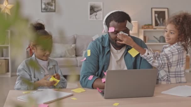 Medium slow shot di due bambine giocose che si divertono a mettere appunti appiccicosi colorati sulla testa e sul corpo dei loro padri mentre lui lavora su laptop con videoconferenza - Filmati, video