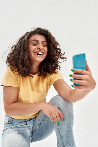 Kıvırcık saçlı neşeli genç bir kadın selfie çekiyor ya da açık arkaplanda izole akıllı telefon kullanarak görüntülü görüşme yapıyor. - Fotoğraf, Görsel