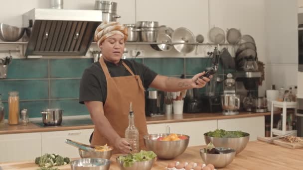 Medium of young African American woman wearing turban and apron, standing at table in professional kitchen, holding smartphone on tripod, showing food and ingredients on camera - Footage, Video
