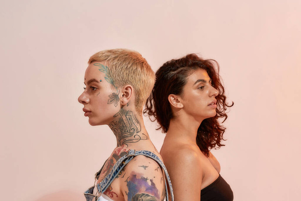 Portrait of two young diverse women, generation Z lesbian couple standing back to back, posing together isolated over light pink background - Fotografie, Obrázek