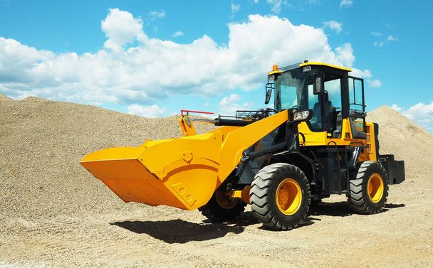 Black-yellow front loader with small wheels against the background of a large pile of stone sand and a blue sky with white clouds.  - Photo, Image