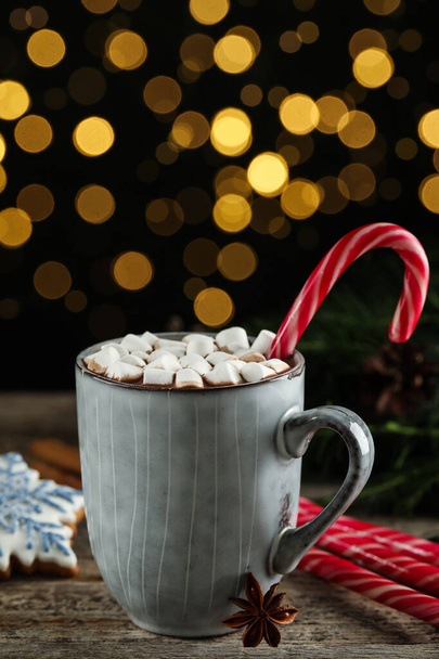 Delicious hot chocolate with marshmallows and candy cane on wooden table against blurred lights - 写真・画像