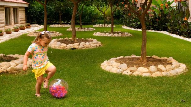 lustige süße kleine Mädchen, 3 Jahre alt, mit zwei Pferdeschwänzen auf dem Kopf, trägt ein buntes T-Shirt und gelbe Shorts, spielt mit einem bunten Ball auf einer grünen Wiese im Garten ihres - Foto, Bild