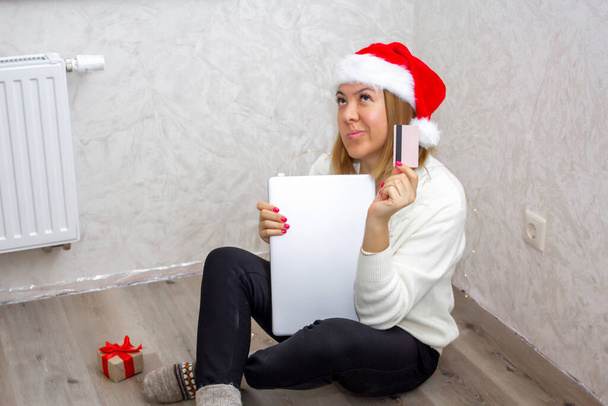 A happy body positive girl in a Santa hat is sitting on the floor of her new empty apartment and choosing gifts for Christmas and New Year online using a laptop. Online shopping of repair goods. - Фото, изображение