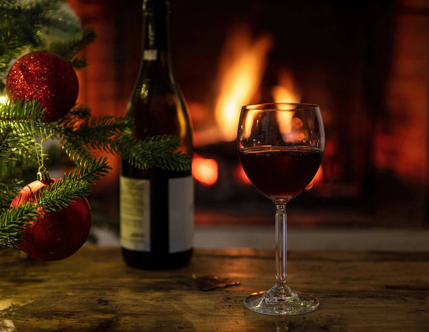  Christmas, red wine glass and bottle on a wooden table, burning fireplace background. Xmas tree decoration. Cozy warm home, winter holidays relaxation - Photo, image