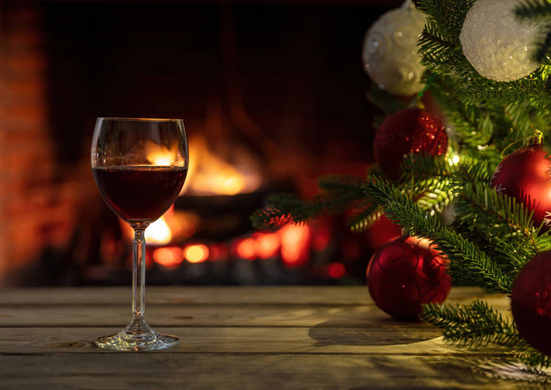  Christmas, red wine glass on a wooden table, burning fireplace background. Xmas tree decoration. Cozy warm home interior, winter holidays relaxation - Фото, изображение