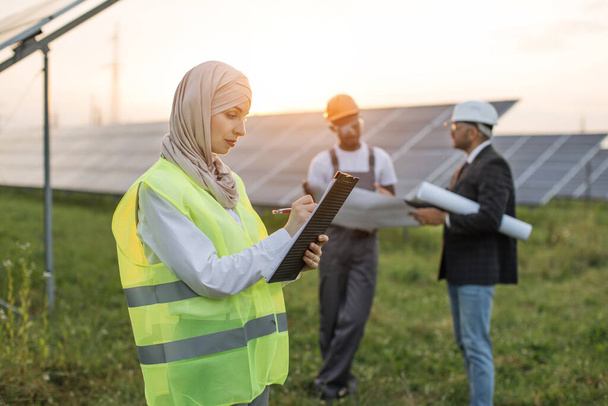 Muslim woman writing on clipboard at solar station - Photo, Image
