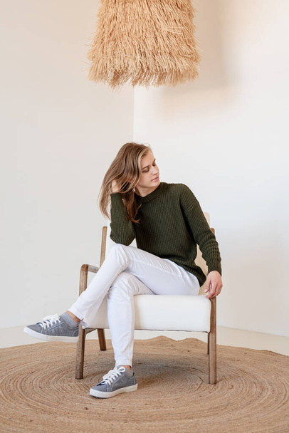 Minimaal licht en luchtig interieur. Aantrekkelijke jonge vrouw in witte jeans en groene sweater zittend op de stoel in licht en luchtig interieur met gesloten ogen - Foto, afbeelding