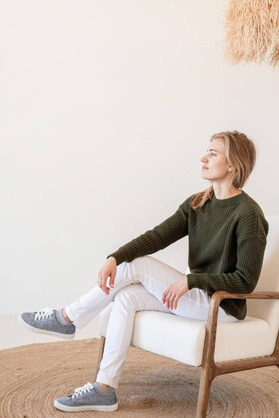 Diseño interior mínimo ligero y aireado. Atractiva joven mujer en jeans blancos y suéter verde sentada en la silla en el interior luminoso y aireado - Foto, imagen