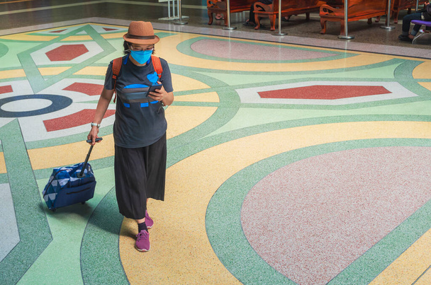 Vista completa de la mujer viajera asiática en máscara protectora usando un teléfono inteligente mientras camina con equipaje en el pavimento en la estación de tren Hua Lamphong en Bangkok, Tailandia - Foto, Imagen