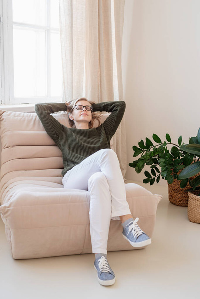 Minimal light and airy interior design. Attractive young woman in white jeans and green sweater sitting on the chair in light and airy interior with plants - Photo, image