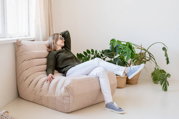 Minimales Licht und luftiges Interieur. Attraktive junge Frau in weißen Jeans und grünem Pullover sitzt auf dem Stuhl im hellen und luftigen Interieur mit Pflanzen - Foto, Bild