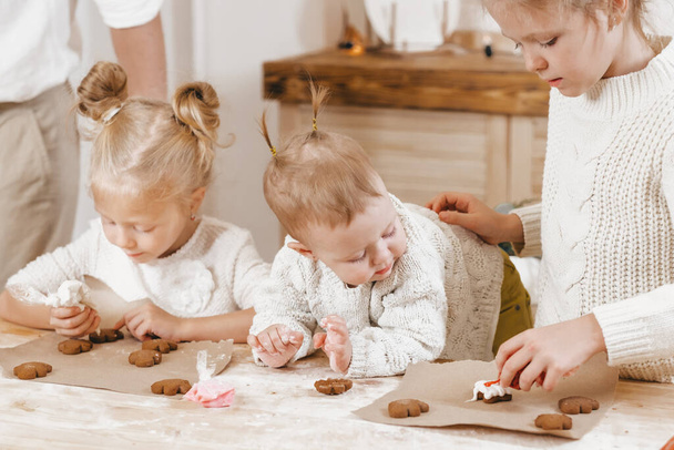 3 meninas loiras decorar biscoitos de Natal com cobertura em uma mesa de madeira na cozinha. As namoradas estão cozinhando juntas na cozinha com decoração de Natal. Relação calorosa de irmãs. Preparação para a mesa de Natal - Foto, Imagem