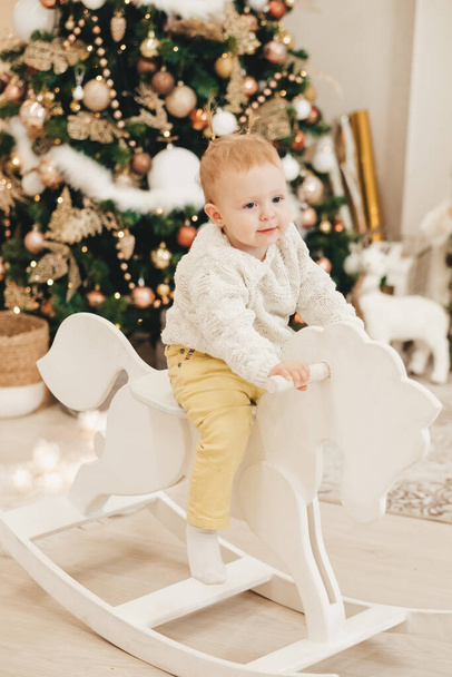 A blonde girl in a sweater rides a wooden horse at home against the background of the Christmas tree. Cute little girl playing at home on the background of garlands. - Foto, Imagem