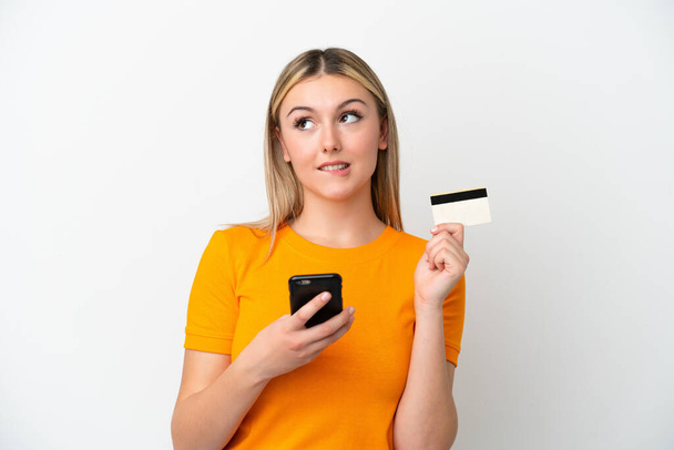 Junge kaukasische Frau isoliert auf weißem Hintergrund kauft mit dem Handy mit einer Kreditkarte, während sie denkt - Foto, Bild